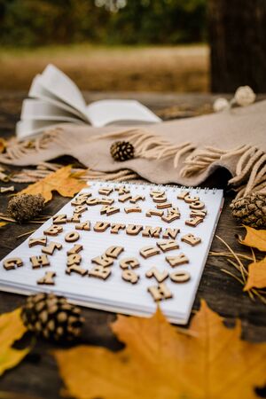 White blank notebook and pen with wooden letters the word autumn on a dark background with a scarf, plaid and a cup, autumn yellow leaves and pine cones around. View from above. flat lay, in blur. Copy spaceの写真素材