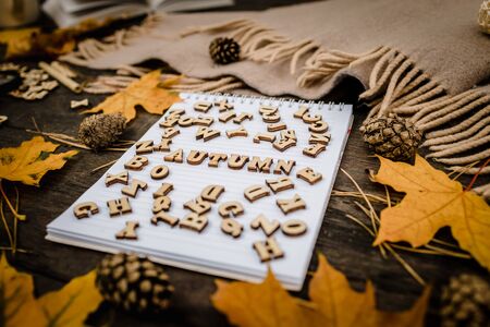 White blank notebook and pen with wooden letters the word autumn on a dark background with a scarf, plaid and a cup, autumn yellow leaves and pine cones around. View from above. flat lay, in blur. Copy spaceの写真素材