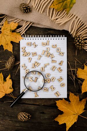 notebook with wooden letters and a magnifying glass on a dark background with a scarf, plaid, around autumn yellow leaves and pine cones. View from above. flat lay. Copy space.の写真素材
