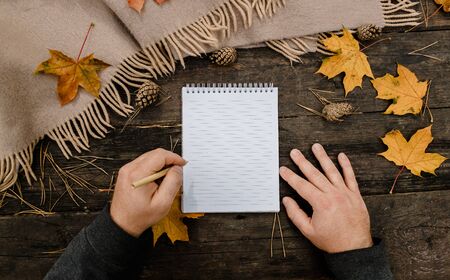 Human hands with a cup of coffee and scarf at wooden table with notebook and pen and autumn leaves. Human hands with a cup of coffee and scarf at wooden tableの写真素材