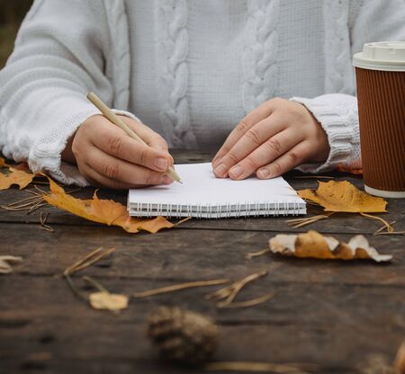 Human hands with a cup of coffee and scarf at wooden table with notebook and pen and autumn leaves. Human hands with a cup of coffee and scarf at wooden tableの写真素材