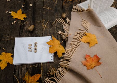 Hands and White blank notebook with wooden letters phrase Good day on a dark background with a scarf, plaid and cup, autumn yellow leaves and pine cones around. View from above. flat lay, in blur. Copy spaceの写真素材