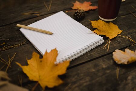 White blank notebook and pen on a dark background with a scarf, plaid and a cup, autumn yellow leaves and pine cones around. View from above. flat lay, in blur. Copy space.の写真素材