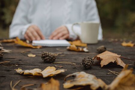 young woman in a white sweater sits at an old dark table in the park, holds a cup of tea with coffee and reads and writes on the background of yellow autumn leaves. Warm autumn atmosphere, soft focus. Copy space.の写真素材