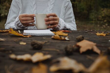 young woman in a white sweater sits at an old dark table in the park, holds a cup of tea with coffee and reads and writes on the background of yellow autumn leaves. Warm autumn atmosphere, soft focus. Copy space.の写真素材