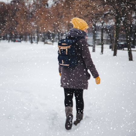 Happy young european woman in a yellow knitted hat and mittens with a backpack, laughs and leaves under the snowfall. View from the back. Retro effect. Blur.の写真素材