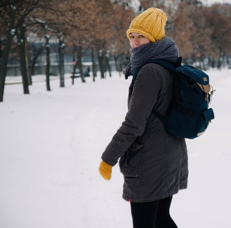 Happy young european woman in a yellow knitted hat and mittens with a backpack, laughs and leaves under the snowfall. View from the back. Retro effect. Blurの写真素材