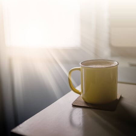 A yellow cup of coffee stands on the office desk in the sunny morning-evening light, next to the monitor and keyboard.の写真素材