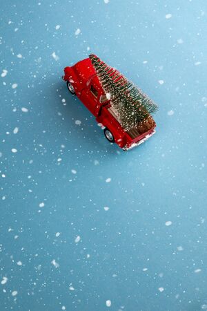 miniature red car in the back carries a Christmas tree on a simple blue-aquamarine background under snow, top view. Save the space. Concept, festive Christmas mood, gift delivery. The trendの写真素材