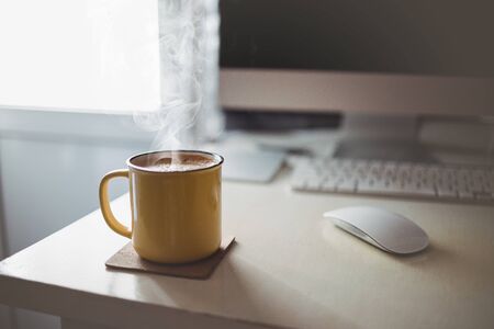 A yellow cup of coffee stands on the office desk in the sunny morning-evening light, next to the monitor and keyboardの写真素材