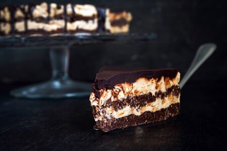 Slice and chocolate cake layers with butter-cream-nut cream with chocolate drips on a glass stand on a dark wooden background. Minimalism, Copy space. Selective focus, close-upの写真素材