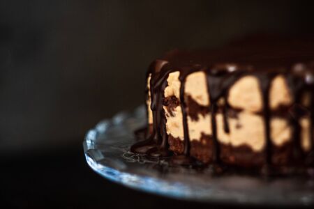 Chocolate round cake layers with butter-cream-nut cream with chocolate drips on a glass stand on a dark wooden background. Minimalism, Copy space. Selective focus, close-up..の写真素材