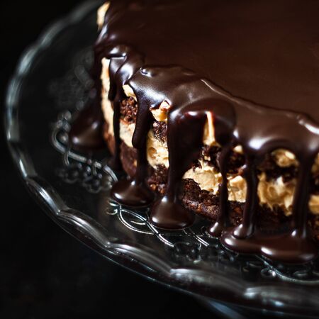 Chocolate round cake layers with butter-cream-nut cream with chocolate drips on a glass stand on a dark wooden background. Minimalism, Copy space. Selective focus, close-up..の写真素材