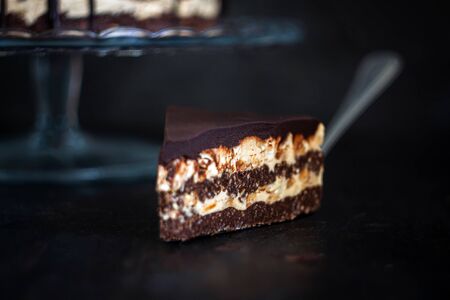 Slice and chocolate cake layers with butter-cream-nut cream with chocolate drips on a glass stand on a dark wooden background. Minimalism, Copy space. Selective focus, close-up.の写真素材