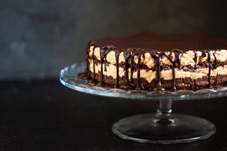 Chocolate round cake layers with butter-cream-nut cream with chocolate drips on a glass stand on a dark wooden background. Minimalism, Copy space. Selective focus, close-up.の写真素材