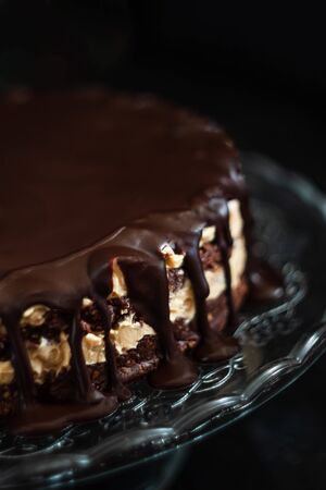 Chocolate round cake layers with butter-cream-nut cream with chocolate drips on a glass stand on a dark wooden background. Minimalism, Copy space. Selective focus, close-up.の写真素材