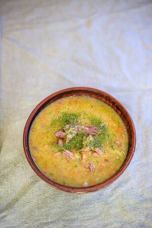 Pea soup with smoked meat, crackers and herbs in a rustic clay plate on a light linen tablecloth. Minimalism Life style, naturalness. Copy space.の写真素材