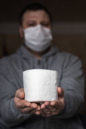 A masked man is holding a roll of toilet paper in his hands. Selective focus. Panic and shortage in stores in the World Pandemic Coronavirus.の写真素材