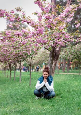 Young happy smiling woman with jeans pants, white sweater and blue scarf sits on the grass under a blossoming pink sakura tree in a park in the city of Kiev, Ukraineの写真素材