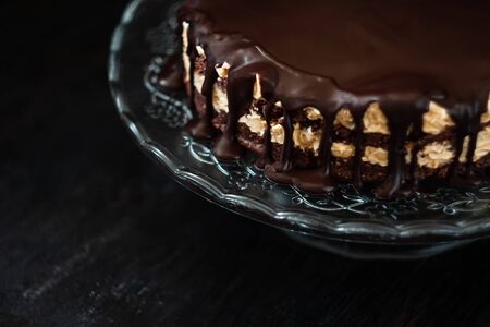 Chocolate round cake layers with butter-cream-nut cream with chocolate drips on a glass stand on a dark wooden background. Minimalism, Copy space. Selective focus, close-up.の写真素材
