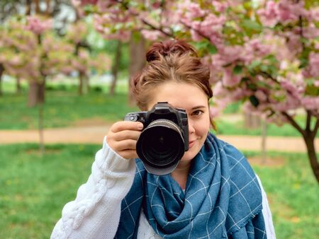 young happy smiling red-haired woman with a white sweater and blue scarf holds in her hands and looks at the camera near a blossoming pink sakura tree in a park in the city of Kiev, Ukraineの写真素材