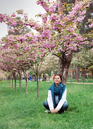 Young happy smiling woman with jeans pants, white sweater and blue scarf sits on the grass under a blossoming pink sakura tree in a park in the city of Kiev, Ukraineの写真素材