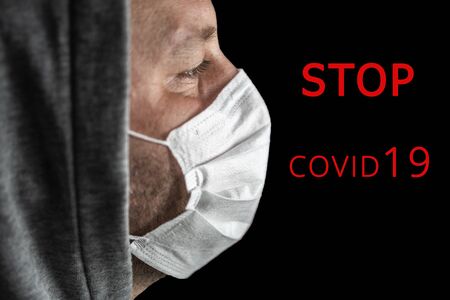 Young tired man in a hood and a medical mask looking forward, red text STOP COVID 19, on a dark background. Copy space. Close-up portrait in profile. Concept, World pandemic coronavirus. Stop the coronavirus epidemic. Health and prevention of influenza and an infectious outbreak.の写真素材