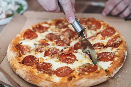 cropped male hand cuts pizza margarita with pepperoni with a special knife, close-upの写真素材
