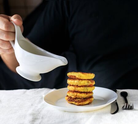 A circumcised man in a black T-shirt holds a white gravy boat in his hands and pours the curd cheesecakes on a plain white tablecloth with sour cream sauce. Concept, simple healthy breakfastの写真素材