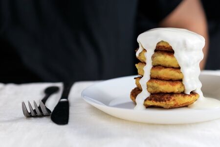 A clipped man in black clothes sits at a table with a linen light tablecloth with curd cheese pancakes in a plate watered with sour cream sauce., Knife, fork. Concept, simple healthy breakfast. Copy space. Side view, selective focus.の写真素材