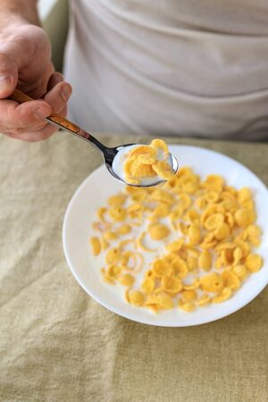 A cropped man holds a spoon in cornflakes with milk in a white plate on a plain rough tablecloth. View from above. Concept, simple fast american healthy breakfast.の写真素材