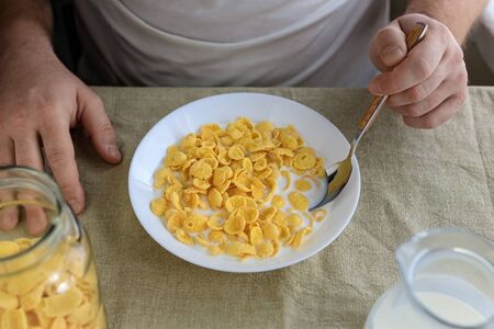 A cropped man holds a spoon in cornflakes with milk in a white plate on a plain rough tablecloth. View from above. Concept, simple fast american healthy breakfast.の写真素材