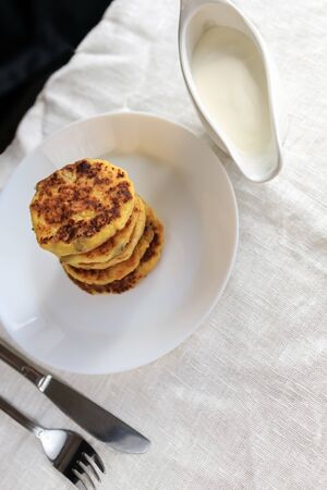 On the table with a linen bright tablecloth there is a white gravy boat with sour cream sauce, curd cheese pancakes in a plate, a fork, a knife on a white plain tablecloth. Concept, simple healthy breakfast. Copy space. View from above. Minimalismの写真素材