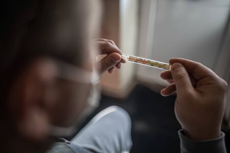 The masked man is looking at a thermometer, a high red temperature of 38 Celsius. Selective focus. Concept, world pandemic of coronavirus Covid-19. Signs of the disease.の写真素材