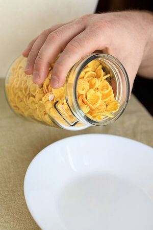 A cropped man sprinkles cornflakes in a white plate on a plain rough tablecloth. Close-up. Top view. Selective focus. Concept, simple fast american healthy breakfastの写真素材