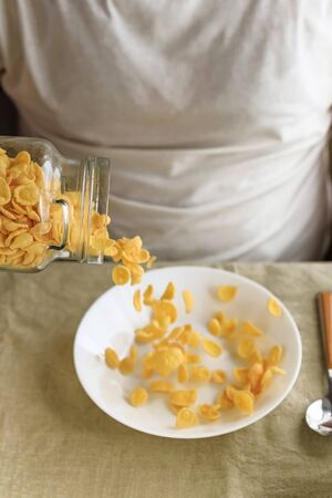 A cropped man sprinkles cornflakes in a white plate on a plain rough tablecloth. Close-up. Top view. Selective focus. Concept, simple fast american healthy breakfast.の写真素材
