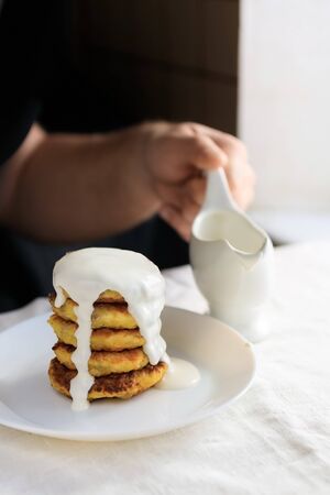 A circumcised man in a black T-shirt holds a white gravy boat in his hands and pours the curd cheesecakes on a plain white tablecloth with sour cream sauce. Concept, simple healthy breakfastの写真素材