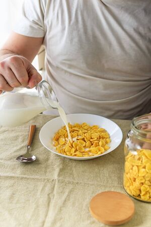 Cropped man pours milk into cornflakes in a white plate on a plain rough tablecloth. View from above. Selective focus. Concept, simple fast american healthy breakfastの写真素材