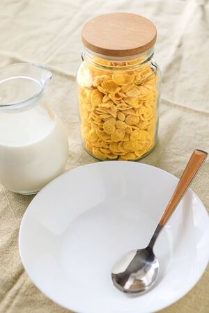 Cornflakes in a jar, next to a transparent jug of milk, an empty plate with a spoon on a plain rough tablecloth. View from above.の写真素材
