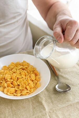 Cropped man pours milk into cornflakes in a white plate on a plain rough tablecloth. View from above. Selective focus. Concept, simple fast american healthy breakfast.の写真素材