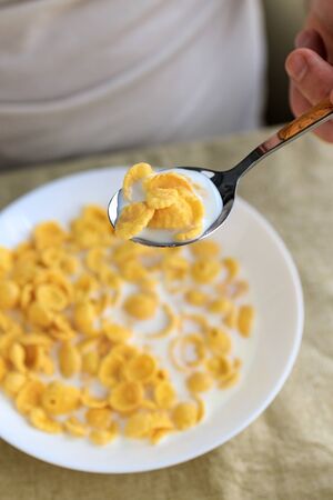 A cropped man holds a spoon in cornflakes with milk in a white plate on a plain rough tablecloth. View from above. Concept, simple fast american healthy breakfast.の写真素材