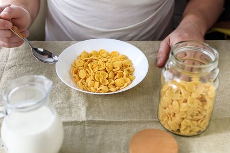 A cropped man holds a spoon in cornflakes with milk in a white plate on a plain rough tablecloth. View from above. Concept, simple fast american healthy breakfast.の写真素材