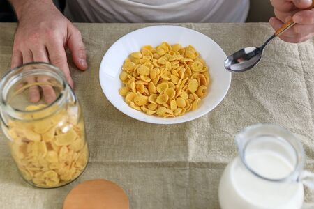 A cropped man holds a spoon in cornflakes with milk in a white plate on a plain rough tablecloth. View from above. Concept, simple fast american healthy breakfastの写真素材
