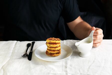 A circumcised man in a black T-shirt holds a white gravy boat in his hands and pours the curd cheesecakes on a plain white tablecloth with sour cream sauce. Concept, simple healthy breakfastの写真素材
