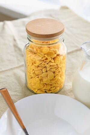 Cornflakes in a jar, next to a transparent jug of milk, an empty plate with a spoon on a plain rough tablecloth. View from aboveの写真素材