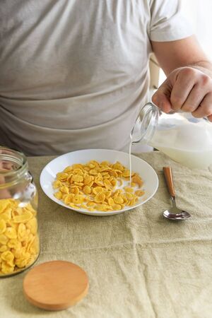 Cropped man pours milk into cornflakes in a white plate on a plain rough tablecloth. View from above. Selective focus. Concept, simple fast american healthy breakfast.の写真素材