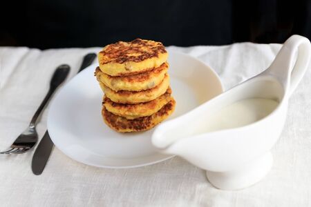 A cropped man in a black T-shirt with a table on which stands a white gravy boat with sour cream sauce, curd cheese pancakes in a plate, fork, knife on a white plain tablecloth. Concept, simple healthy breakfast. Copy space. View from above. Minimalism.の写真素材