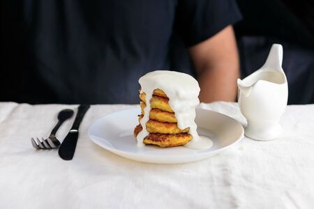 A cropped man in a black T-shirt with a table on which stands a white gravy boat with sour cream sauce, curd cheese pancakes in a plate, fork, knife on a white plain tablecloth. Concept, simple healthy breakfast. Copy space. View from above. Minimalism.の写真素材