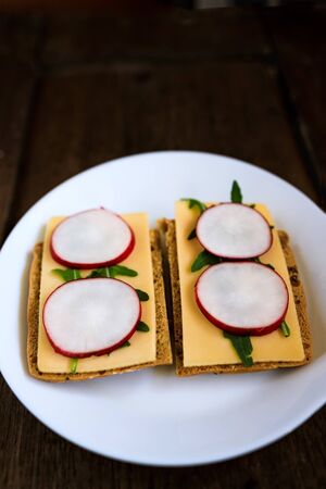 Sandwich made from rye bread, hard cheese, radish, arugula, served on a white plate against a dark wooden background. Symmetry. Copy space. Flat lay, top view. Close-upの写真素材