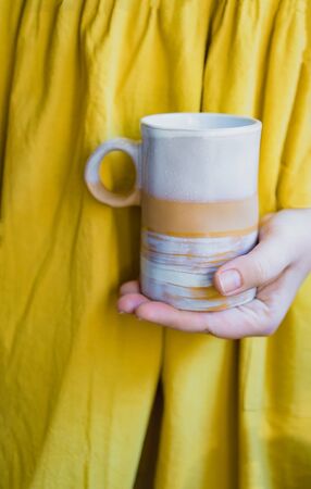 Cropped young white woman in a simple yellow linen dress holds in her hands a clay ceramic cup of an original handmade shape. Copy space. Trendの写真素材